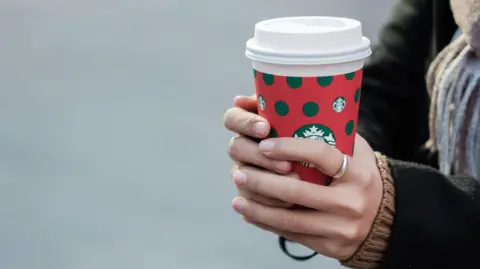 Getty Images A woman holds a Christmas-themed Starbucks coffee cup. She's wearing a black coat, scarf and a silver ring on her left index finger.
