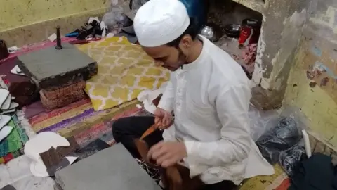 A worker sits cross-legged on the floor making shoes